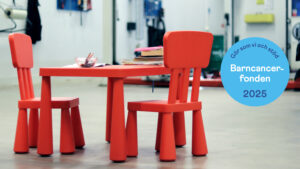 Ett rött pysselbord för barn står i en besiktningshall med pysselböcker och en pennburk på. Till höger en illustration/logga med Barncancerfondens budskap.